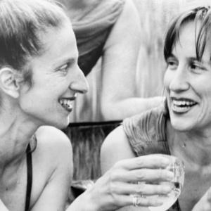 Juliet Palmer & Karen Kaeja smile, holding wineglasses, wearing cocktail dresses while sitting in a fountain.