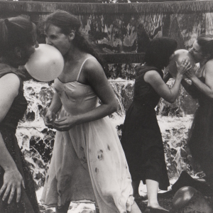 black & white photo: 5 dancers in cocktail dresses in a fountain hold balloons between them in pairs using their mouths to hold them.