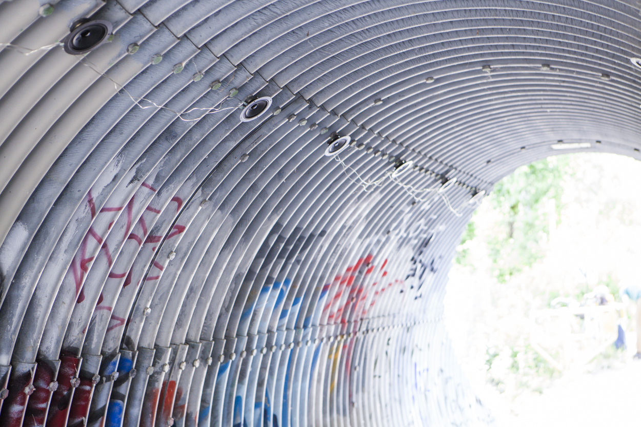Inside a galvanized metal tunnel with graffiti and small speakers attached at intervals