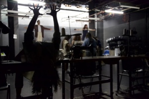 Photograph of opera singers in a set that resembles a sewing sweatshop. There are sewing machines and fluorescent lights. One singer is arched backwards over a sewing table, her hands raised to the ceiling. 