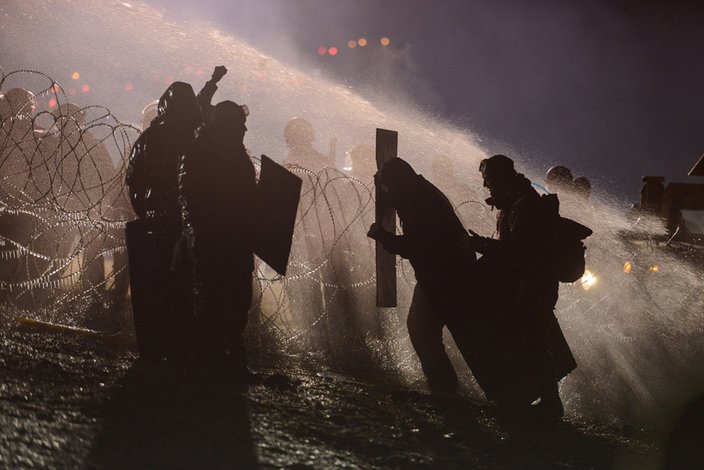 Water protectors with improvised shields at night silhouetted by a powerful light lean into the force of a water cannon. They are separated from helmeted police officers by a tangle of barbed wire