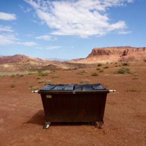 A large black dumpster sits in the desert of the Southwest, a mesa in the background and blue sky with a smattering of light clouds